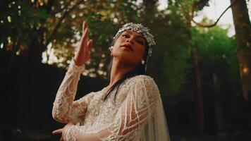 Elegant woman in vintage bridal attire with headpiece, posing in a garden setting at dusk. video