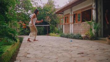 femme en marchant avec une arrosage pouvez près une traditionnel maison avec luxuriant verdure. video