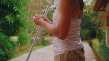 Close-up of a woman in casual clothing holding a wooden stick, walking in a lush garden pathway. video