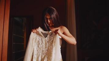 Smiling bride in a dimly lit room holding up her elegant lace wedding dress, preparing for her special day video