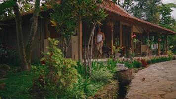 tranquillo scena di un' persona in piedi su il veranda di un' rustico Casa circondato di lussureggiante verdura e un' pietra percorso. video