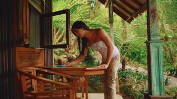 sereno tropical configuração com uma mulher limpeza a de madeira mesa, cercado de exuberante vegetação. video