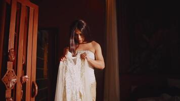 Elegant woman in a strapless dress holding a lace gown, contemplating it thoughtfully in a warmly lit room video