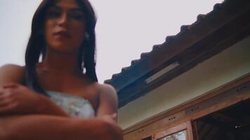 kant visie van een attent vrouw in een wit jurk staand door een rustiek huis, omarmen haarzelf met een sereen uitdrukking video