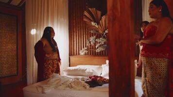 dos mujer en tradicional atuendo en un dormitorio, uno en pie por un cama y el otro por un ventana, en calentar Encendiendo. video