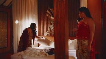 zwei Frauen im traditionell Kleidung im ein Schlafzimmer, einer Stehen durch ein Bett und das andere durch ein Fenster, im warm Beleuchtung. video