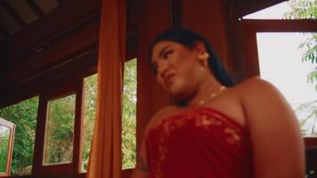 Person in a wig and dress sitting indoors, looking at a red fabric with a thoughtful expression. video