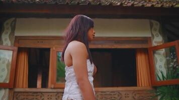 Side profile of a young woman with long hair standing in front of a rustic house with traditional architectural video