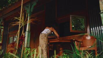 Person leaning on a wooden railing of a rustic house surrounded by lush greenery. video