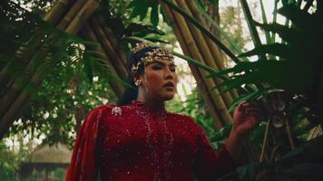 Majestic woman in red traditional attire with headpiece, posing in a lush greenhouse setting. video