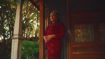 Elegant woman in red dress standing by wooden house amidst lush greenery video