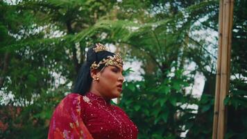 traditionell asiatisk kvinna i kulturell klädsel med huvudstycke stående i en frodig trädgård. video
