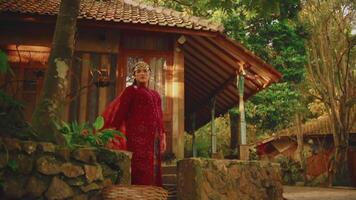 persona en rojo túnica caminando por tradicional casa rodeado por lozano verdor. video