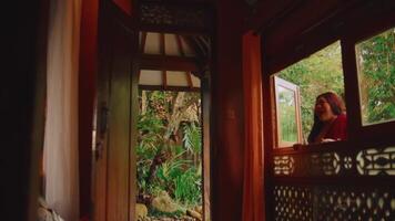 Tranquil scene of a woman gazing out from a traditional wooden window, overlooking a serene garden. video