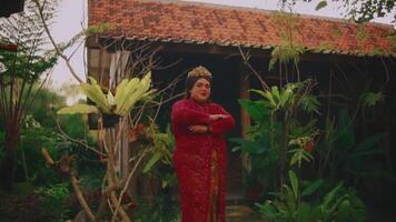 kvinna i traditionell röd klänning stående i främre av en rustik hus med grönska. video