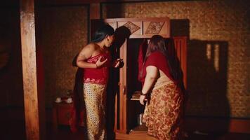 twee Dames in traditioneel zuiden Aziatisch kleding innemend in een cultureel ceremonie binnenshuis, met een vrouw aanpassen de anderen kleding video