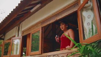 Frau im rot Kleid genießen das Aussicht von ein rustikal Kabine Fenster, umgeben durch Natur. video