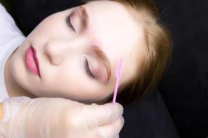 a close-up of the face of a blonde model who lies on the eyebrow lamination procedure, the master combs the eyebrows with a brush photo