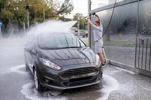 The man in the background has raised the water cannon above his head and is aiming it at the roof of the auto. A car at a self service car wash. photo