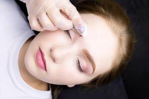 a close-up of the face of a blonde model who is lying on a black couch, the master wipes excess paint from her eyebrows with a cotton sponge photo