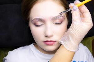 the blonde is lying on the procedure of long-term eyebrow styling, the master applies laminating compositions with a brush to the eyebrows photo