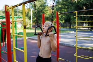 A boy with an athletic build pulls himself up on the suspended handrails. Street workout on a horizontal bar in the school park. photo