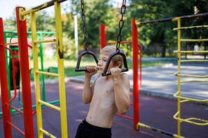 A boy with an athletic build pulls himself up on hanging rails. Street workout on a horizontal bar in the school park. photo