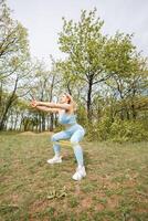 Girl athlete training in the park exercises gluteal muscles do squats. Beautiful blonde Caucasian woman in blue tight tracksuit. Blonde girl at an outdoor training session photo