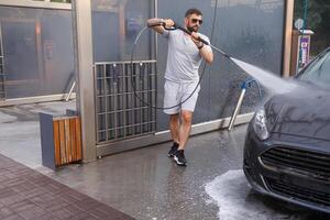 un hombre con un agua cañón en su mano carreras alrededor el coche y lavados él. un coche a un yo Servicio coche lavar. foto