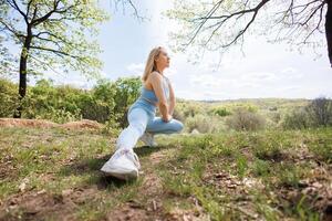 The girl is doing a stretching squat on one leg. Beautiful blonde Caucasian woman in blue tight tracksuit. Blonde girl at an outdoor training session photo