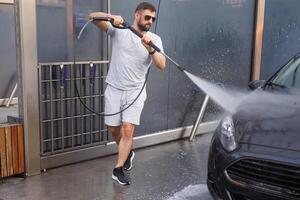 un hombre con un agua cañón en su mano carreras alrededor el coche y lavados él. un coche a un yo Servicio coche lavar. foto