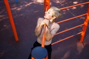 Athletic child performs a lateral pull-up on the bar. Street workout on a horizontal bar in the school park. photo