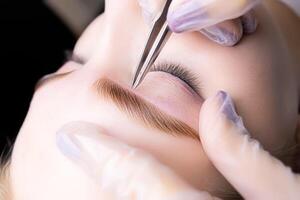 a close-up of the eyebrows of the model, the master holds in his hand a tweezer that removes excess hairs of already colored and laminated eyebrows photo