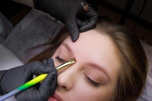 Using a brush, the master applies marking lines on the model's eyebrows before permanent eyebrow makeup. PMU Procedure, Permanent Eyebrow Makeup. photo