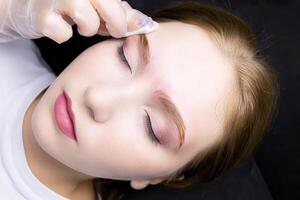 the girl model at the eyebrow lamination procedure, the master erases the remnants of their laminating compositions with a cotton sponge photo