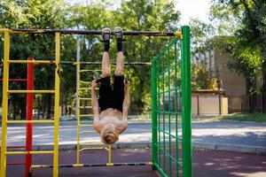 The boy hung on the sports rings upside down, holding them with his hands. Street workout on a horizontal bar in the school park. photo