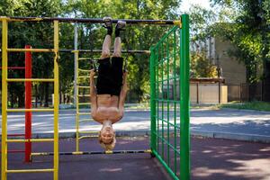 The boy hung upside down on the sports rings with his hands on them. Street workout on a horizontal bar in the school park. photo