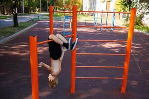 a teenager of an athletic build hangs head down, clinging to the horizontal bar with his feet. Street workout on a horizontal bar in the school park. photo