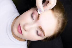 a close-up of the face of a blonde model who is lying on a black couch, the master wipes excess paint from her eyebrows with a cotton sponge photo