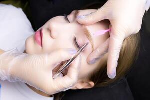 model during the eyebrow lamination procedure, the master plucks out the excess hairs in the eyebrows with tweezers after the lamination procedure photo