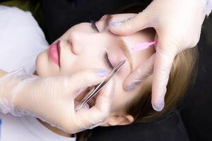 model during the eyebrow lamination procedure, the master plucks out the excess hairs in the eyebrows with tweezers after the lamination procedure photo