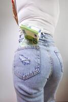 The girl stands back and has a hundred euro bill sticking out of the back pocket of her jeans. A girl in jeans with a euro banknote in her hands. photo
