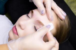the girl model at the eyebrow lamination procedure, the master erases the remnants of their laminating compositions with a cotton sponge photo