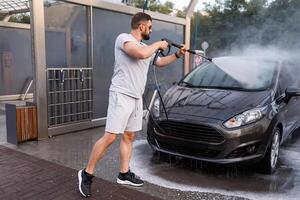 un hombre soportes en frente de el coche y puntos un agua cañón a él, Lavado el agua apagado de él. un coche a un yo Servicio coche lavar. foto