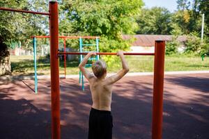 The teenager stands with his back holding on to the sports bar. Street workout on a horizontal bar in the school park. photo