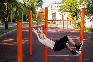 un atlético adolescente se balancea en diferente direcciones, colgando en el horizontal bar. calle rutina de ejercicio en un horizontal bar en el colegio parque. foto