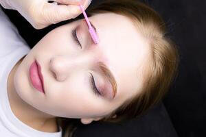 a close-up of the face of a blonde model who lies on the eyebrow lamination procedure, the master combs the eyebrows with a brush photo