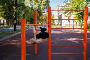 un atléticamente construido adolescente es balanceo en diferente direcciones mientras colgando en un bar. calle rutina de ejercicio en un horizontal bar en el colegio parque. foto