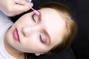 a close-up of the face of a blonde model who lies on the eyebrow lamination procedure, the master combs the eyebrows with a brush photo