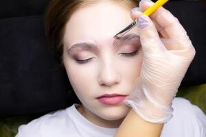the blonde is lying on the procedure of long-term eyebrow styling, the master applies laminating compositions with a brush to the eyebrows photo